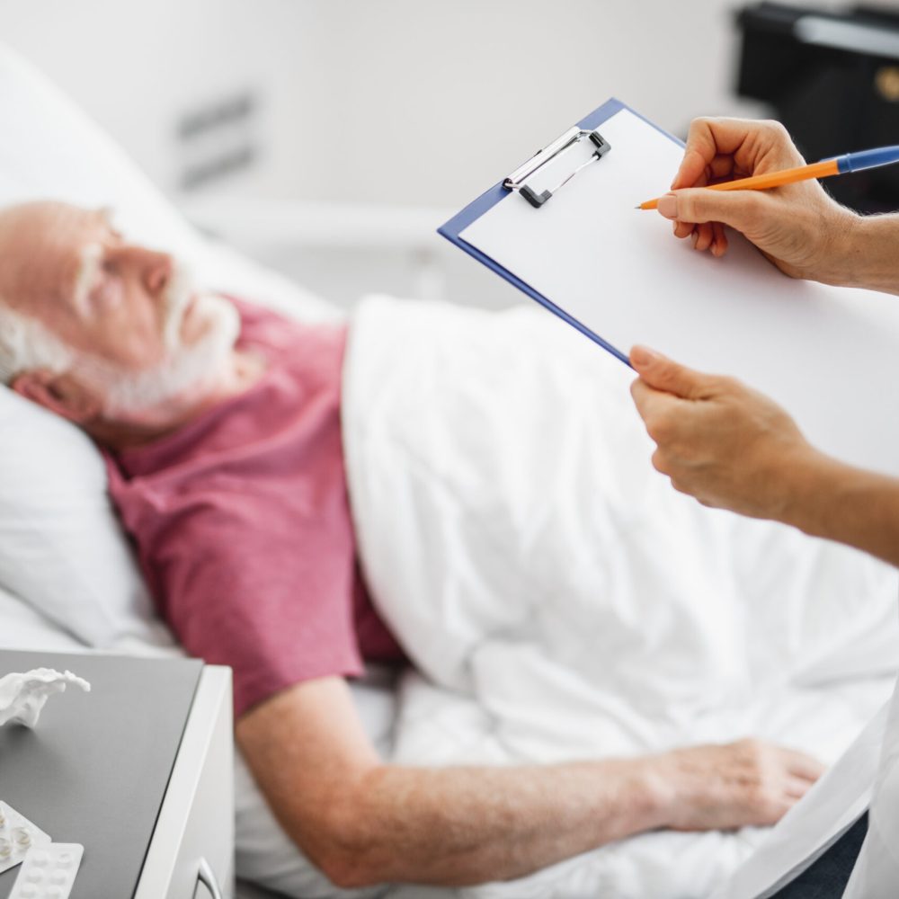 Female therapist filling up medical form while standing near nightstand with medications in hospital room. Old man sleeping on blurred background